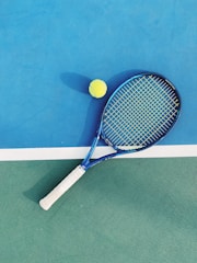 A tennis racket with a blue frame and white grip lies on a two-toned tennis court. Next to it is a yellow tennis ball. The court features a vibrant blue section and a green section divided by a white line.