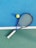 A tennis racket with a blue frame and white grip lies on a two-toned tennis court. Next to it is a yellow tennis ball. The court features a vibrant blue section and a green section divided by a white line.