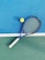 A tennis racket with a blue frame and white grip lies on a two-toned tennis court. Next to it is a yellow tennis ball. The court features a vibrant blue section and a green section divided by a white line.