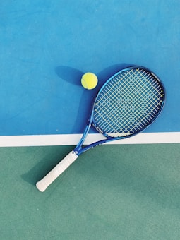A tennis racket with a blue frame and white grip lies on a two-toned tennis court. Next to it is a yellow tennis ball. The court features a vibrant blue section and a green section divided by a white line.