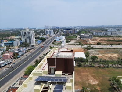 Aerial view of an urban area with a main road running through the center. On the left side, there are several buildings, including a tall apartment complex. The right side shows open plots of land with sparse vegetation. The sky is overcast, and there appear to be vehicles on the road. On the rooftop of the nearest building, solar panels are installed.