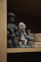 Three small, clay figurine sculptures are displayed on a wooden shelf. The figurines appear to be handcrafted and represent different characters, two of whom seem to be wearing accessories or distinctive clothing. There are also other clay objects visible in the background.