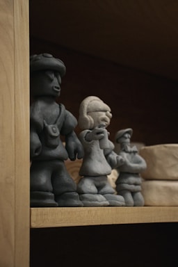 Three small, clay figurine sculptures are displayed on a wooden shelf. The figurines appear to be handcrafted and represent different characters, two of whom seem to be wearing accessories or distinctive clothing. There are also other clay objects visible in the background.
