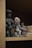 Three small, clay figurine sculptures are displayed on a wooden shelf. The figurines appear to be handcrafted and represent different characters, two of whom seem to be wearing accessories or distinctive clothing. There are also other clay objects visible in the background.