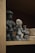 Three small, clay figurine sculptures are displayed on a wooden shelf. The figurines appear to be handcrafted and represent different characters, two of whom seem to be wearing accessories or distinctive clothing. There are also other clay objects visible in the background.