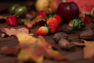 An assortment of autumn-themed items is scattered on a wooden surface, including colorful strawberries, autumn leaves, walnuts, an apple, and a glimpse of a watermelon.