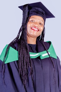 A person is wearing a graduation cap and gown, featuring green accents. The individual is smiling broadly, suggesting a moment of celebration, possibly at a graduation ceremony.