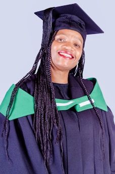 A person is wearing a graduation cap and gown, featuring green accents. The individual is smiling broadly, suggesting a moment of celebration, possibly at a graduation ceremony.
