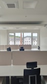 An empty office space with multiple workstations, each equipped with black office chairs and light-colored dividers. Large windows at the far end of the room show an outside view of buildings and a chimney, with a backdrop of a cloudy sky.