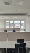 An empty office space with multiple workstations, each equipped with black office chairs and light-colored dividers. Large windows at the far end of the room show an outside view of buildings and a chimney, with a backdrop of a cloudy sky.