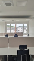 An empty office space with multiple workstations, each equipped with black office chairs and light-colored dividers. Large windows at the far end of the room show an outside view of buildings and a chimney, with a backdrop of a cloudy sky.