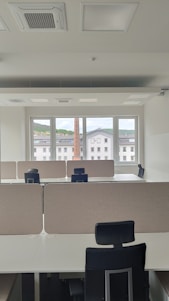 An empty office space with multiple workstations, each equipped with black office chairs and light-colored dividers. Large windows at the far end of the room show an outside view of buildings and a chimney, with a backdrop of a cloudy sky.