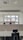 An empty office space with multiple workstations, each equipped with black office chairs and light-colored dividers. Large windows at the far end of the room show an outside view of buildings and a chimney, with a backdrop of a cloudy sky.