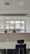 An empty office space with multiple workstations, each equipped with black office chairs and light-colored dividers. Large windows at the far end of the room show an outside view of buildings and a chimney, with a backdrop of a cloudy sky.