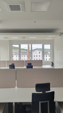An empty office space with multiple workstations, each equipped with black office chairs and light-colored dividers. Large windows at the far end of the room show an outside view of buildings and a chimney, with a backdrop of a cloudy sky.