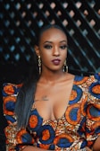 A woman with dark hair styled straight wears colorful, patterned clothing featuring circular designs in orange, blue, and white. She has bold makeup with dark red lipstick and eyeliner, and wears large, gold hoop earrings. The background is a dimly lit with a geometric pattern.