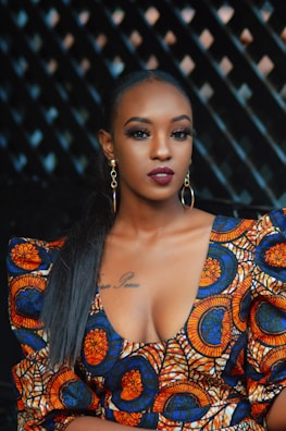 A woman with dark hair styled straight wears colorful, patterned clothing featuring circular designs in orange, blue, and white. She has bold makeup with dark red lipstick and eyeliner, and wears large, gold hoop earrings. The background is a dimly lit with a geometric pattern.