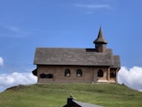 A small, rustic chapel with a wooden exterior sits atop a grassy hill. The structure features a steeply pitched roof and a single, pointed steeple. Its windows are arched, and the building is set against a backdrop of a clear blue sky with a few scattered clouds.