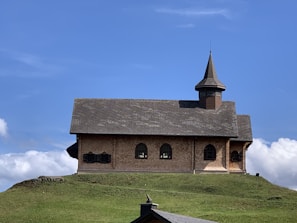 A small, rustic chapel with a wooden exterior sits atop a grassy hill. The structure features a steeply pitched roof and a single, pointed steeple. Its windows are arched, and the building is set against a backdrop of a clear blue sky with a few scattered clouds.