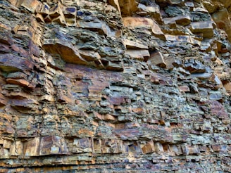 A close-up view of a rock formation with rugged, stratified layers in shades of brown, gray, and tan. The texture is rough and uneven, showcasing natural geological patterns and erosion.