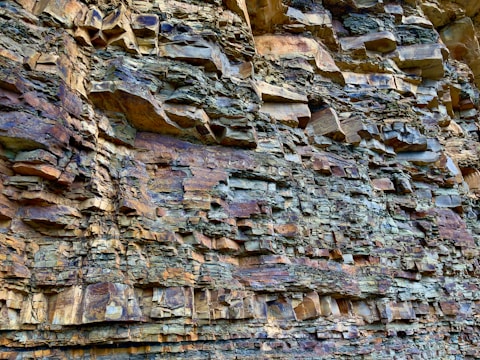 A close-up view of a rock formation with rugged, stratified layers in shades of brown, gray, and tan. The texture is rough and uneven, showcasing natural geological patterns and erosion.