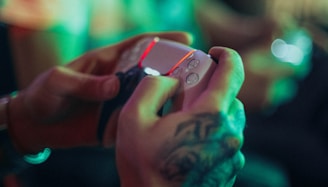 A person with a tattoo on their hand is holding a white gaming controller. The ambient lighting features green and red hues, creating a vibrant atmosphere.