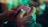 A person with a tattoo on their hand is holding a white gaming controller. The ambient lighting features green and red hues, creating a vibrant atmosphere.