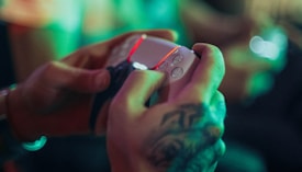 A person with a tattoo on their hand is holding a white gaming controller. The ambient lighting features green and red hues, creating a vibrant atmosphere.