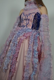 A person wearing an ornate gown featuring flowing ruffled details in pastel pink, lavender, and blue hues. The dress has intricate patterns and layers, with semi-transparent fabric on the sleeves and a creatively designed bodice.