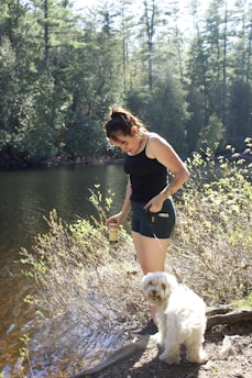 A woman in a black tank top and shorts standing next to a river or lake, holding a can in her left hand while looking down. She is accompanied by a small white fluffy dog on a leash. The surrounding area is densely wooded with tall trees and thick vegetation, and the ground near the water is covered with bushes and small plants.