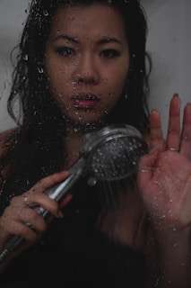 A person holds a showerhead close to their face, with water droplets visible on a glass surface in front of them. The lighting is dim, casting a moody atmosphere, and the person appears to have wet hair, with a contemplative or serious expression.