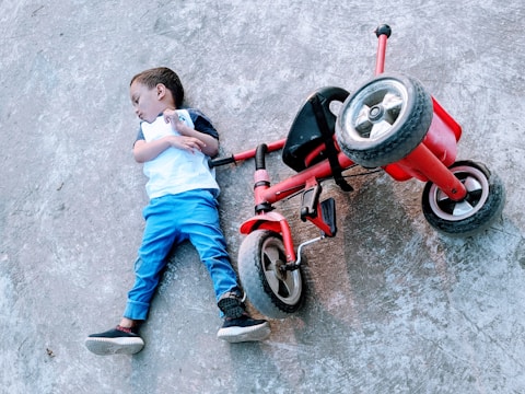 A child is lying on the pavement next to a fallen red tricycle. The child appears to be wearing a white shirt and blue pants.