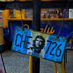 A vintage license plate featuring an image of Che Guevara is hanging on a chain in front of a food truck with a bright blue and yellow exterior. There are drinks and a menu visible inside the truck.