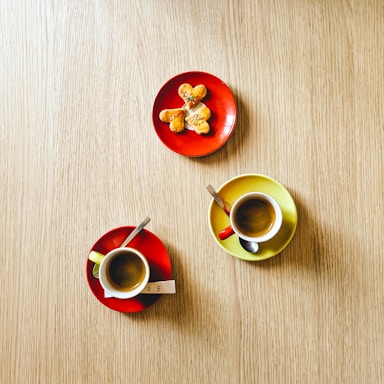A wooden table displays two cups of espresso on colorful saucers. One cup is on a red saucer with a sugar packet labeled 'RE' and a spoon, while the other is on a yellow saucer with a spoon. Above them, a red plate holds four heart-shaped cookies arranged in a star pattern, topped with small colorful sprinkles.