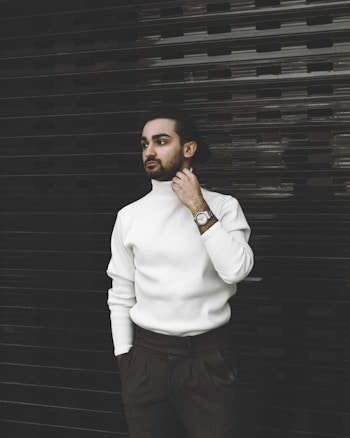 A person with a contemplative expression stands against a dark, textured background. They are wearing a white turtleneck sweater and dark pants. The individual has a watch on their wrist and one hand is near their neck, adding a sense of style and elegance.