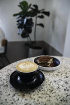 A cappuccino with intricate latte art is served in a black cup and saucer on a speckled granite table beside a small dessert, likely tiramisu or cake. In the background, a potted green plant adds a touch of nature to the indoor setting, and part of a chair can be seen to the left.