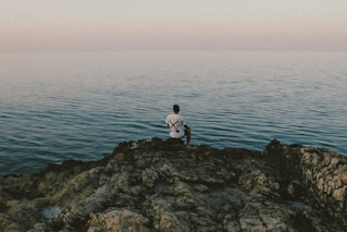 man in white shirt sitting on rock by the sea during daytime