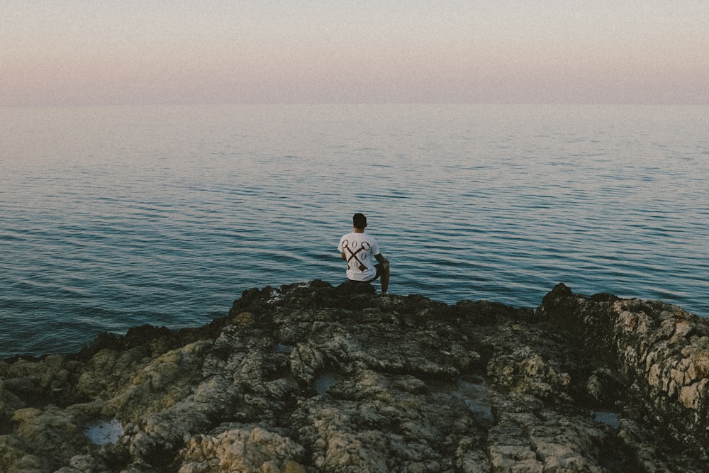 man in white shirt sitting on rock by the sea during daytime