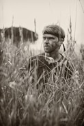 A person with a headband and camouflage paint on their face is partially concealed amidst tall grasses, evoking a sense of stealth or concealment. The setting suggests a natural or outdoor environment, possibly for activities like hunting or military maneuvers.