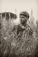 A person with a headband and camouflage paint on their face is partially concealed amidst tall grasses, evoking a sense of stealth or concealment. The setting suggests a natural or outdoor environment, possibly for activities like hunting or military maneuvers.