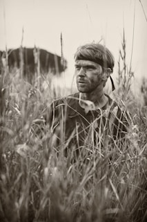 A person with a headband and camouflage paint on their face is partially concealed amidst tall grasses, evoking a sense of stealth or concealment. The setting suggests a natural or outdoor environment, possibly for activities like hunting or military maneuvers.