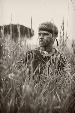 A person with a headband and camouflage paint on their face is partially concealed amidst tall grasses, evoking a sense of stealth or concealment. The setting suggests a natural or outdoor environment, possibly for activities like hunting or military maneuvers.
