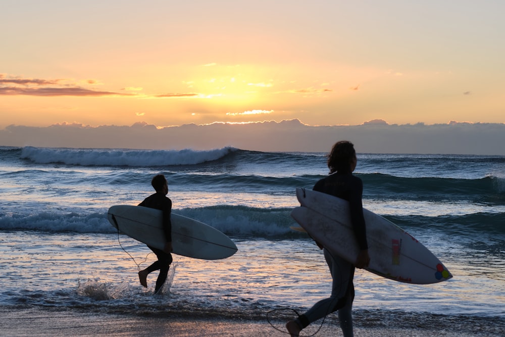 Hombre sosteniendo tabla de surf blanca caminando en la playa durante la puesta del sol