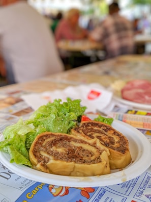 A plate with two slices of savory roll filled with meat, served with a side of fresh green lettuce, placed on a table. The background depicts a blurred setting, seemingly a busy indoor place with people seated and engaging in activities.