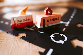 Two toy vehicles are positioned on a track, one is a red and white ice cream truck with a cone on top, while the other is an orange donut truck with a donut figure on top. The track is black with white road markings, including a circular roundabout symbol. The surface appears to be a wooden floor, suggesting a playful or imaginative environment.