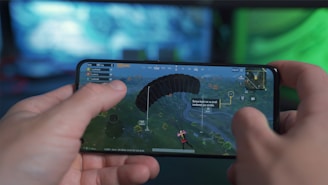 A person is holding a smartphone horizontally, playing a video game that involves a character parachuting into an open area. The screen shows various game interface elements including a compass, a mini-map, and player stats. The background has a blurred and colorful setting, suggesting a focus on the gaming experience.