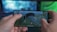 A person is holding a smartphone horizontally, playing a video game that involves a character parachuting into an open area. The screen shows various game interface elements including a compass, a mini-map, and player stats. The background has a blurred and colorful setting, suggesting a focus on the gaming experience.