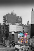The image features an urban cityscape with a large billboard displaying colorful graffiti art. The graffiti showcases expressive, vibrant portraits composed of geometric shapes, creating a sharp contrast with the monochrome buildings surrounding it. In the foreground, there are various vehicles, including cars and a bus, navigating through the streets.