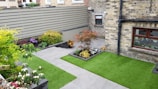 A small, neatly landscaped backyard features green artificial grass and several flower beds with various plants and colorful flowers. The area is enclosed by a brick wall and a wooden fence, providing privacy. A gray stone pathway divides the garden, leading to a door and windows on a brick house facade.