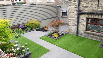 A small, neatly landscaped backyard features green artificial grass and several flower beds with various plants and colorful flowers. The area is enclosed by a brick wall and a wooden fence, providing privacy. A gray stone pathway divides the garden, leading to a door and windows on a brick house facade.